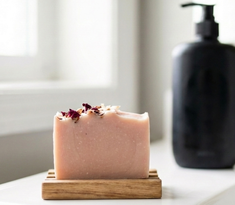 A handcrafted pink coconut and rose soap bar resting on a wooden draining dish in the foreground, contrasted against a blurred, generic black plastic body wash bottle in the background. A visual comparison of eco-friendly natural bar soap versus synthetic liquid detergent